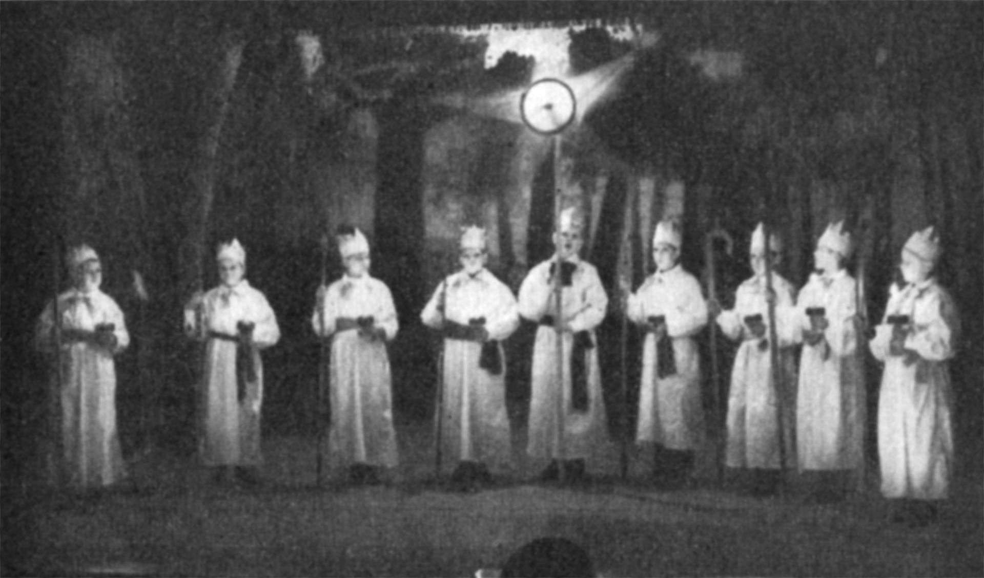 På bilden syns stärngossa i rad med långa vita klänningar och höga hattar på en mörk scen. Bilden är svartvit och från år 1936.