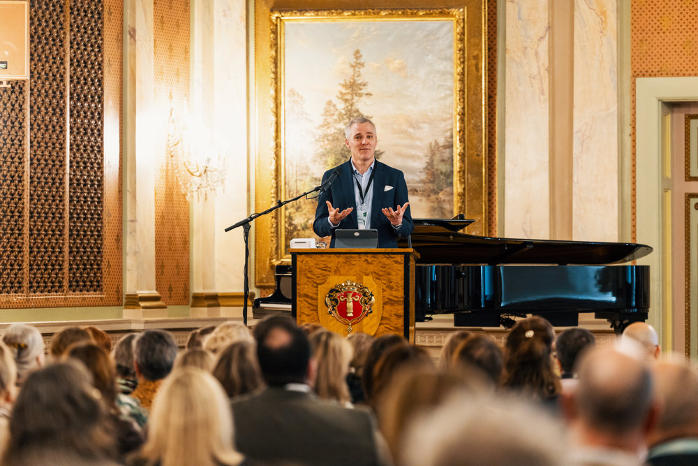 På bilden syns utbildningsminister Anders Adlercreutz, klädd i mörk kostym och ljus skjorta. Han står i ett talarbås i Vasa stadshus på Utbildningskonferensen 2026. Framför honom syns flera rader av mä'nniskor som lyssnar. Bakom honom finns utsmyckade väggar med tapeter och en stor tavla.