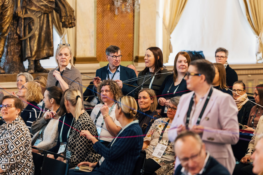 På bilden syns ett tiotal människor i en utsmyckad konferenslokal. En  del står och andra sitter. Runt omkring dem och i deras händer går garntrådar här och tvärs. Foto: Christoffer Björklund