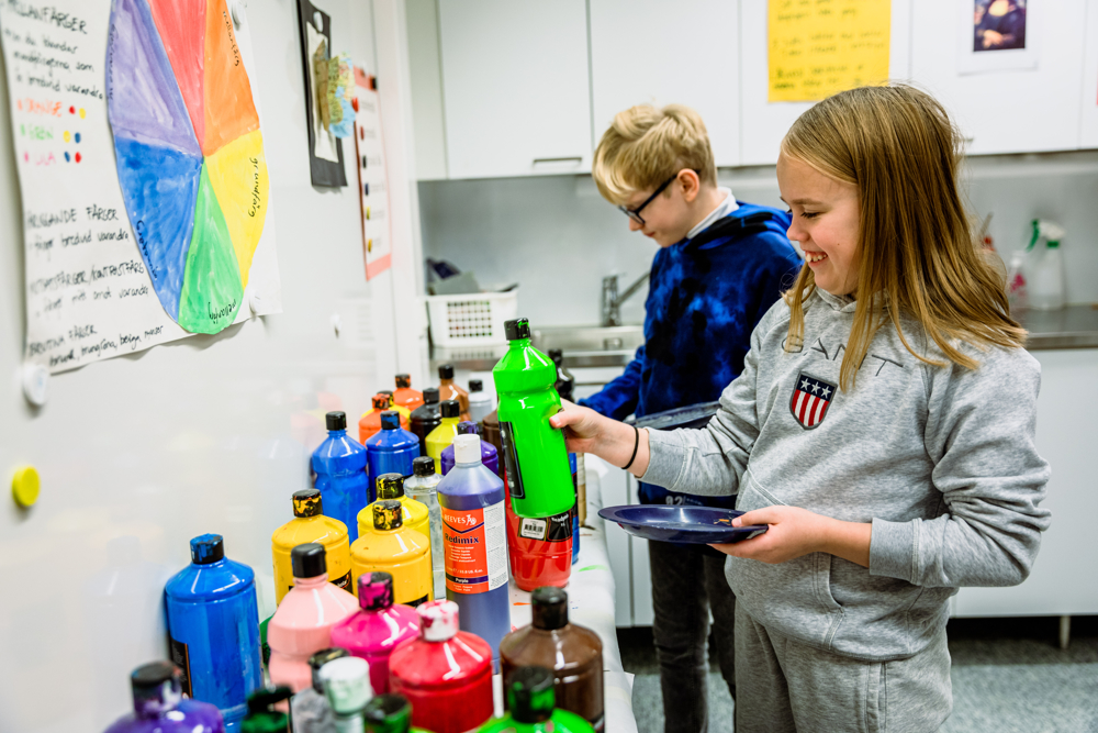 På bilden syns två skolbarn som står vid en bänk med stora färgburkar. Flickan är klädd i grå tröja med tryck på, hon har mellanbrunt hår och står med en burk grön färg i handen och ser glad ut. Man ser hennes tänder när hon ler. Lite längre bort står en blond pojke med blå tröja och glasögon. Han står och väljer färgburk. De har båda blåa små tallrikar i händerna. Framför dem finns en vägg med en färgkarta och i bakgrunden ser man vita klassrumsväggar med planscher på.