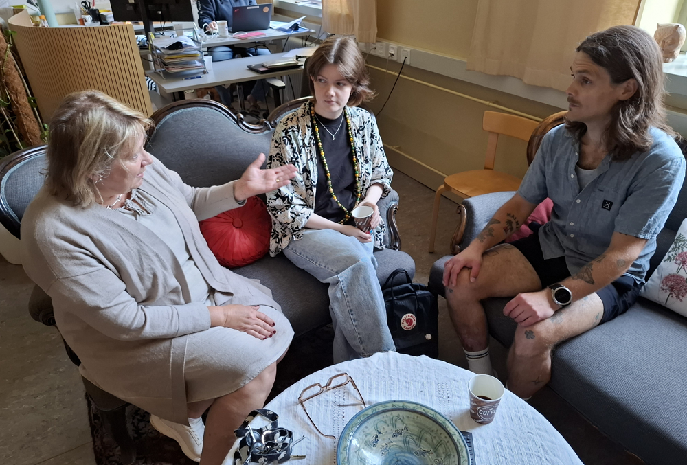 På bilden syns en kvinna i beige jacka och kjol som sitter i en blå soffa tillsammans med en ung kvinna med mörkt hår. Hon har en mönstrad skjortblus ovanpå en svart t-tröja. De samtalar med en ung man som sitter på en annan likadan soffa till höger. Han har kortärmad grå blus och brunt axellångt hår.