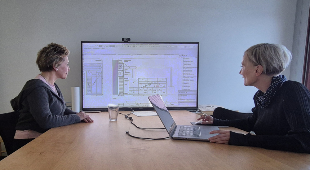På bilden syns två kvinnor, Johanna Stenman, bildkonstlärare, till vänster och inredningsarkitekt Päivi Meuronen till höger. De sitter vid ett ljusbrunt bord och tittar mot en skärm i ändan av bordet. På skärmen ser man en ritning av ett rum och text. Kvinnan till vänster har kort mörkt hår med ljusa slingor i. Hon har en mötkgrå huvtröja med en rosa blus under. Kvinnan till höger har en mörkbrun tröja och en vitprickig scarf runt halsen.
