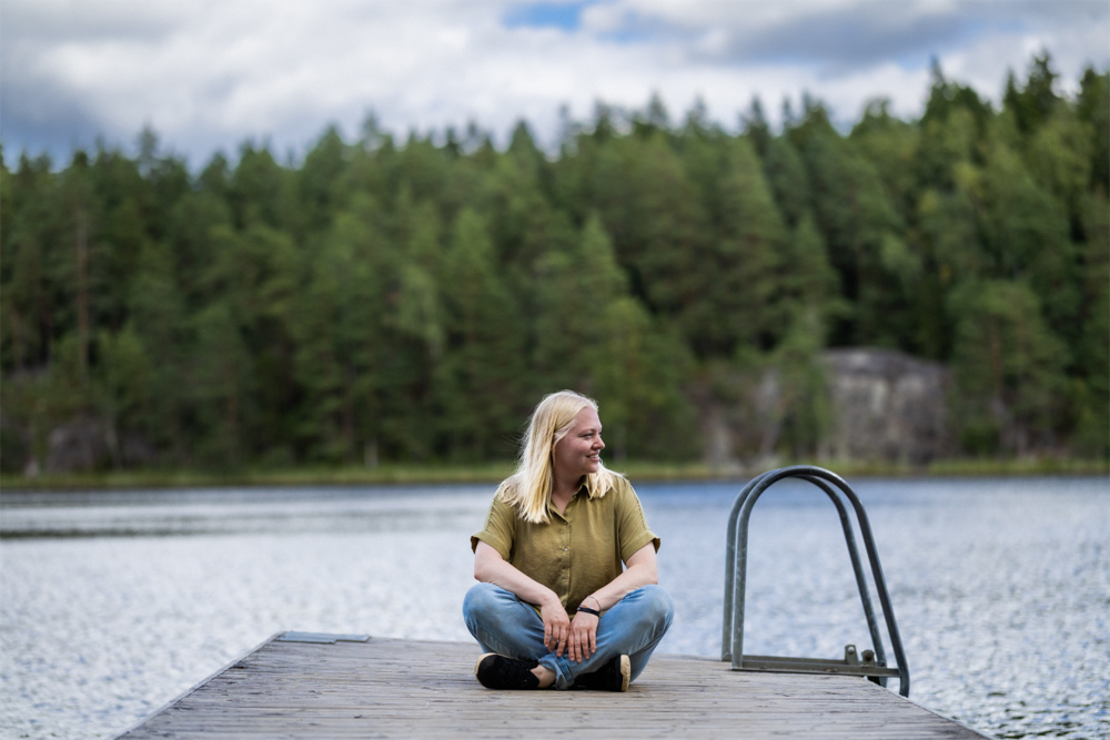 På bilden syns en kvinna i trettioårsåldern som sitter på en brygga med benen i kors. I bakgrunden ser man en sjö och skog. Kvinnan ser lite till höger i bilden. Hon har blont halvlångt hår, en grön bljus och jeans. Hennes namn är Tarja Krum ovh hon får Fredrika Runeberg-stipendiet 2026.