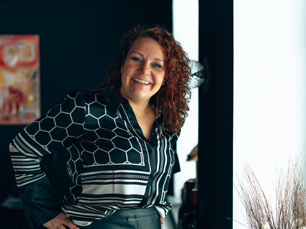 På bilden syns en medelålders kvinna vid namn Anna Häggblom som drar kurser i AI. HOn har lockigt axellångt rött hår, tittar rakt in i kameran och skrattar stort. Hon är klädd i en mörkgrön och vit skjortsblus och står med ena handen på höften. I bakgrunden ser man dels ett fönster med växter utanför, dels en mörk vägg där man blurrad kan skymta en tavla i rött och gult.