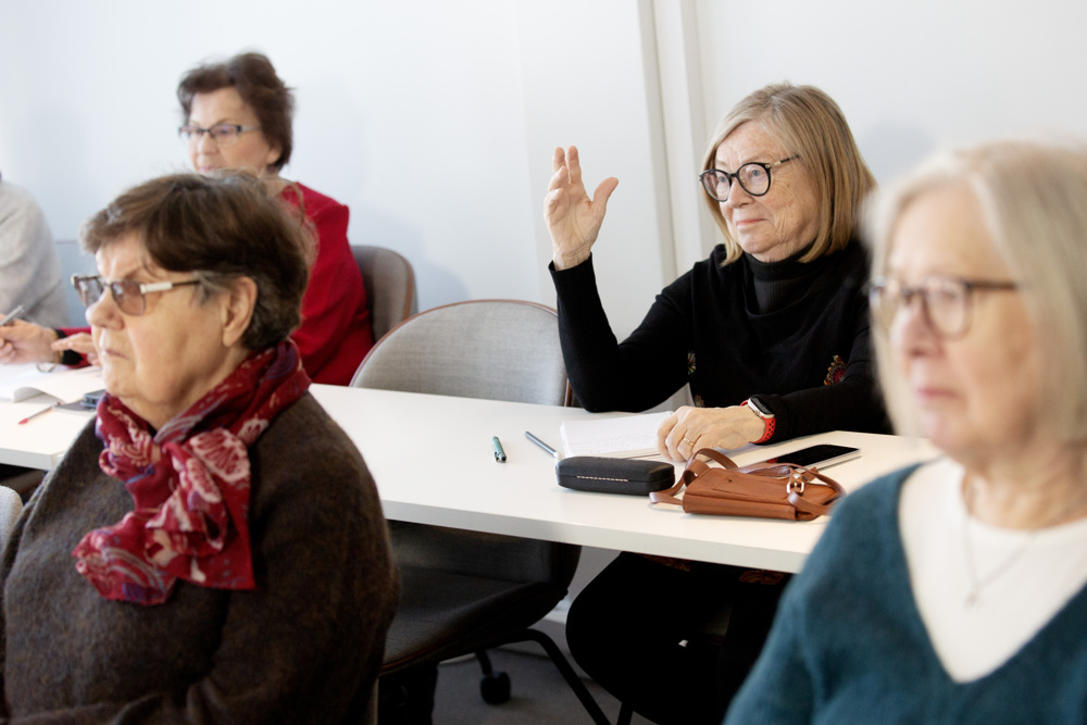 På bilden syns fyra damer i pensionsålder som sitter vid vita bord och har blicken riktad mot någon som talar utanför bild. Bara en av damerna är skarp. Hon sitter och markerar. HOn har svart tröja och under bordet ser man hennes svarta byxor. Hon har pageklippt ljust hår och runda mörka glasögon. Hon ser glad ut. Bakgrunden är en vit vägg.