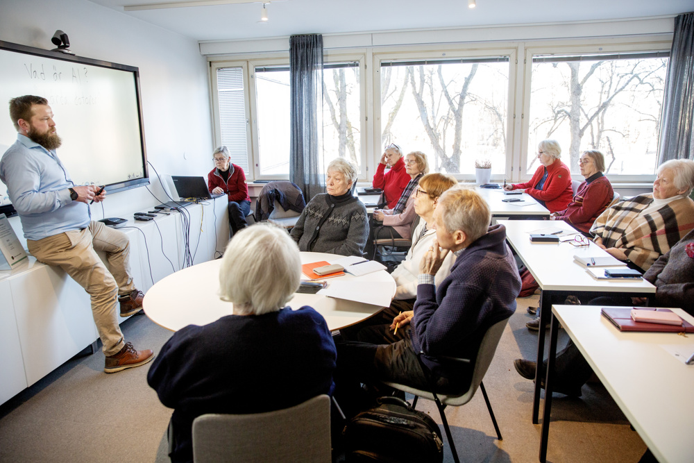 På bilden ses ett rum där det pågår undervisning, om AI för seniorer. Längt till vänster syns en medelåldersman med skägg och brunt hår. Han är klädd i ljusblå skjorta och ljusbruna byxor. Han halvsitter framför en whiteboard. Framför honom vid olika slags vita bord sitter ett flertal personer i pensionsålder och lyssnar. De flesta är kvinnor. I bakgrunden ser man fönster, med kala träd utanför.