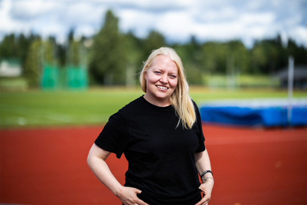 På bilden syns en kvinna i 30-årsåldern, Tarja Krum. Hon har blont halvlångt hår och tittar rakt in i kameran. HOn är klädd i en svart t-shirt och står med händerna i jeansens fickor. I bakgrunden ser man suddigt en idrottsplan med röda löpbanor.