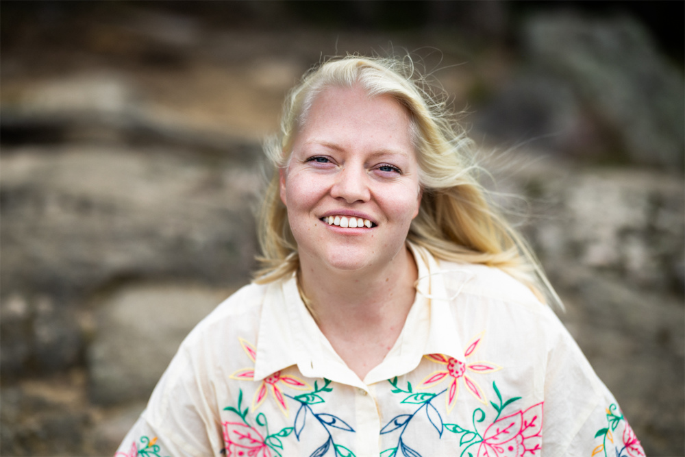På bilden syns kvinna i trettioårsåldern vid namn Tarja Krum. HOn han blont halvlångt hår och är klädd i en vit skjorta med brodier i rosa, orange och grönt. Hon är fotad utomhus och i bakgrunden ser man suddigt växtlighet.
