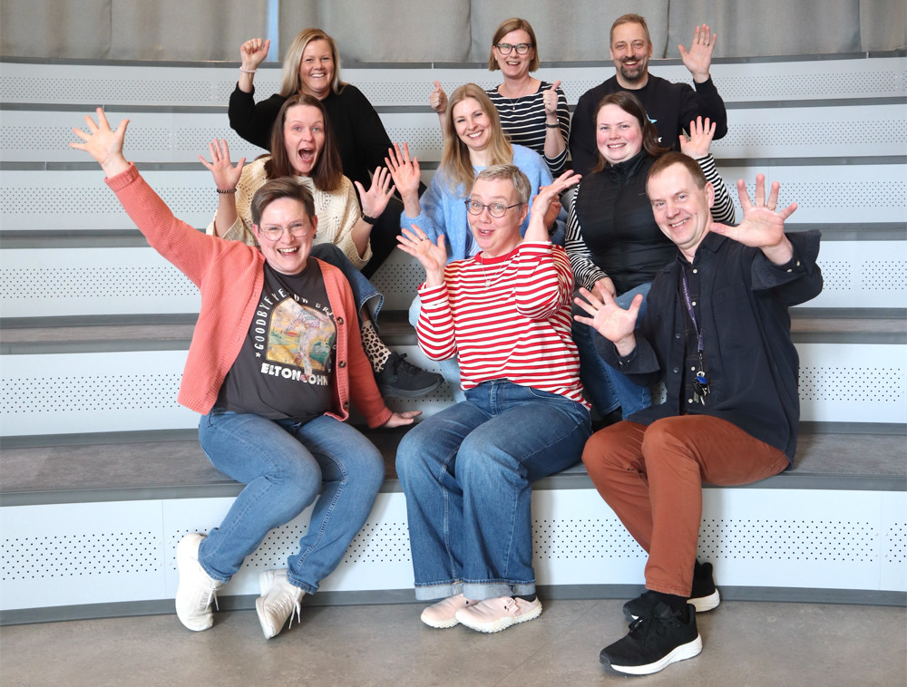 På bilden syns nio personer som arbetar på Zachariasskolan i Nykarleby. De sitter på vita och gråa bänkar i något som ser ut som ett auditorium. De ser alla glada ut och tittar rakt in i kameran. De har alla händerna uppsträckta och en del ser ut att skrika. Två av personerna är män, resten är kvinnor.