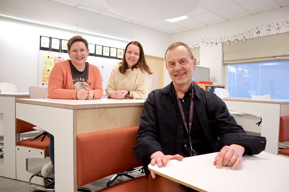 På bilden syns tre personer som sitter i skolbänkar i ett ljust klassrum med vita väggar och vitt tak. Högt uppe på väggen hänger en rad vimplar i klara färger. Längst till vänster i övre raden sitter en kvinna med kort brunt hår och tunna metallbågade glasögon. Hon är klädd i jeans, brun t-tröja med tryck framtill och rosa tröja. Kvinnan till höger har halvlångt brunt hår som är löst, ljus stickad tröja och jeans. Mannen längst till höger som sitter i raden framför dem har kort ljust hår, Mörkblå skjortblus som är uppknäppt ovanpå en mörkblå t-shirt. Han har orangea byxor och en guldring på ett finger. De tittar alla in i kameran och ler stort.