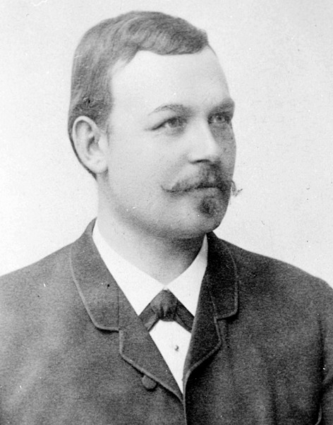 Bilden är en svartvit historisk bild av en ung man med bakåtkammat mörkt hår. Han är klädd i kostym och skjorta och tittar ut tmot höger. Bilden är tagen från sidan.