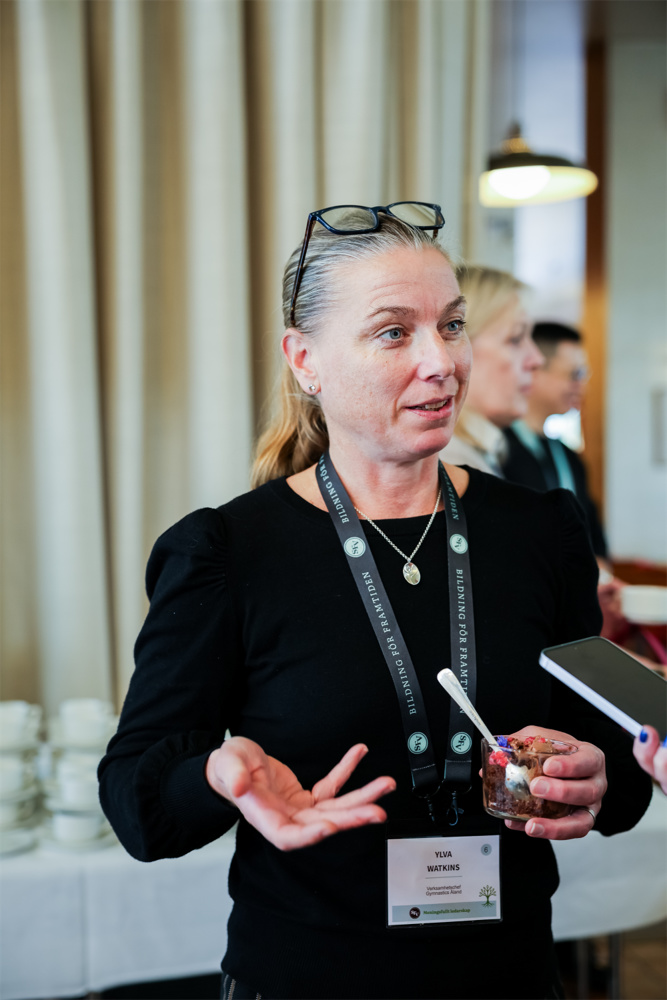 På bilden ser man en kvinna i yngre medelåldern, Ylva Watkins. Hon är klädd i en svart blus och har ett band runt halsen med en namnlapp. HOn har mörkt hår som är draget tillbaka från ansiktet och ett par glasögon på hjässan. Bakgrunden är ett mötesrum med vita gardiner på väggarba.