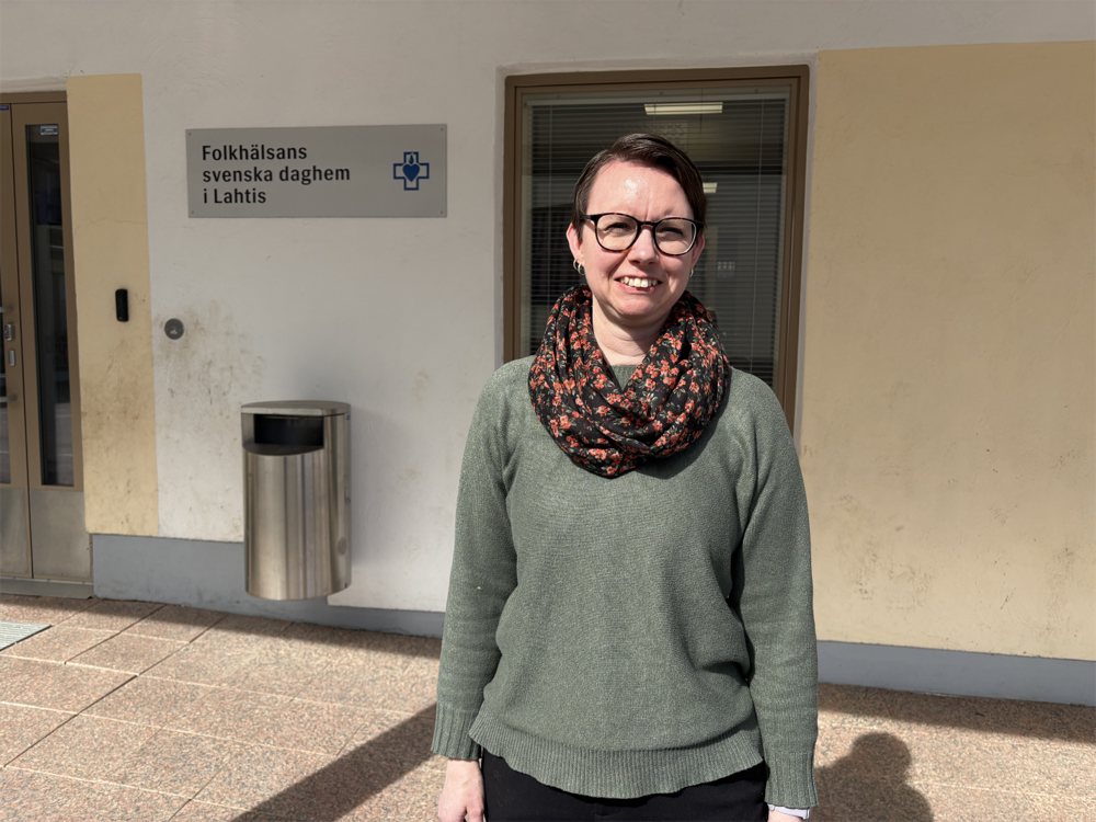 På bilden syns en kvinna med kort mörkt hår och glasögon med mörka bågar. Hon ser allvarlig ut och tittar rakt in i kameran. Hon är klädd i en ljusgrön tröja och har en scarf i svarta med röttt möster runt halsen, Hon ahr mörka byxor och bilden av skuran vid låren. Hons tår framför en byggnad i aprikosfärg och vitt och på en skylt står det Folkhälsans svenska daghem i Lahtis.