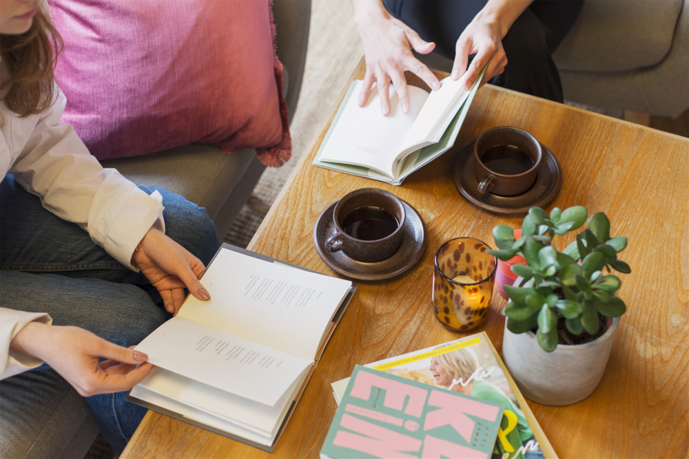 Till höger på bilden syns en yngre kvinna. Man ser en liten del av hennes ansikte, men främst händerna som håller i en bok, och att hon sitter på en soffa iklädd jeans och en vit tröja. Soffan har ett knallrosa överdrag. Till höger om den unga kvinnan ser man en del av en annan kvinnamen mörka byxor. Hon håller också ci en bok som ligger på ett mellanbrunt soffbord mellan dem.På borde syns också värmeljus i glasbehållare och bruna kaffekoppar med fat.