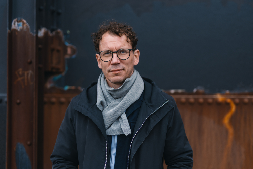 På bilden syns en medelåders man vid namn Juha Itkonen. Han är fotograferad utomhus, han står framför en vägg  i rostbrunt och mörkblått. Han är klädd i en mörkblå jacka som är uppknäppt och runt halsen har han en ljusgrå halsduk. Han har brunt lockigt hår och glasögon. Han ser rakt in i kameran och ser allvarlig ut.