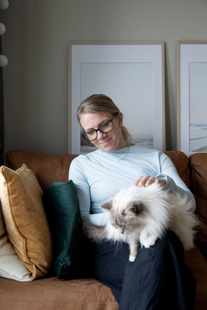  På bilden syns en kvinna vid namn Linda Mannila. Hon leder en kurs om AI. Hon har ljust hår som är draget tillbaka i en hästsvans, hon är klädd i en ljusblå blus och mörka byxor. Hon har glasögon. HOn tittar lite nedåt mot en vit långhårig katt i som står i hennes famn. Hon sitter i en brun soffa med en gul, en grön och en vit soffkudda till vänster. Bakom henne syns en ljusgrön vägg och två tavlor i ljusa färger i tunna träramar.