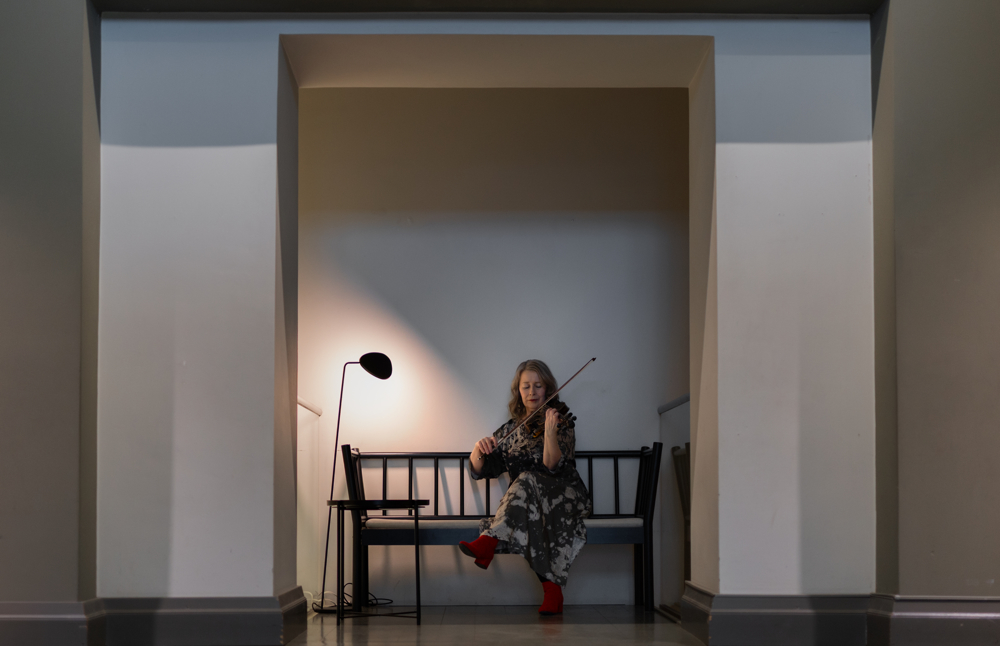 På bilden syns en medelålderns kvinna som sitter på en bänk och spelar fiol. Bilden har väldigt myckwet miljö runt henne hon sitter i en fördjupning i en naturvit och blå vähh. Man ser en lampa bredvid henne och hon sitter på en träbänk med ett ryggstöd av pinnar. HOn har en klänning i gråa nyanser och röda stövletter.