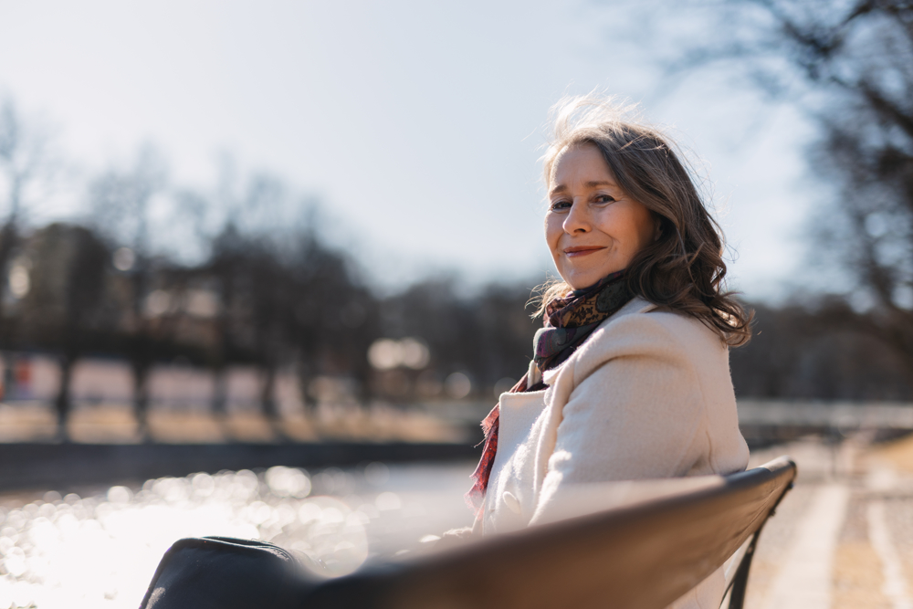 PÅ bilden syns en medelålderns kvinna som sitter på en parkbänk framför en å. I bakgrunden ser man suddigt åns vatten, kala träd och blå himmel. Kvinnann  är klädd i en naturvit yllejacka och har en scarf i blått och brunt runt halsen. Hon har böjligt grått och brunt hår som är ungefär axellångt. HOn tittar in i kameran över en ena axel, fotografen står bakom bänken, och ser glad ut.