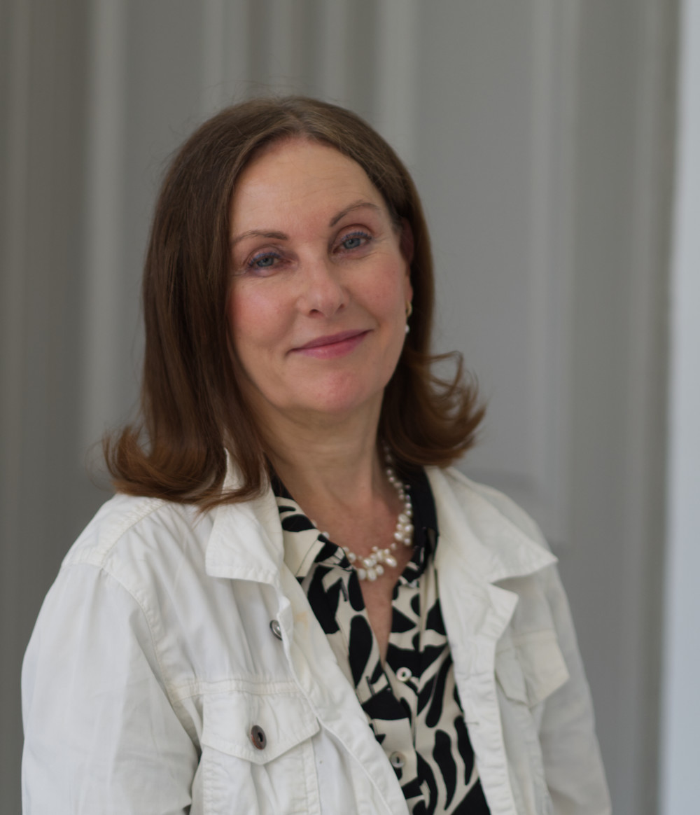 På bilden syns en kvinna i vit jacka och med enmönstrad blus i svart och vitt under. Hon har halvlångt mörkt hår som lockar sig i topparna. Hon ser rakt in i kameran och ser allvarlig ut. I bakgrunden ser man en vit gardin som veckar sig.