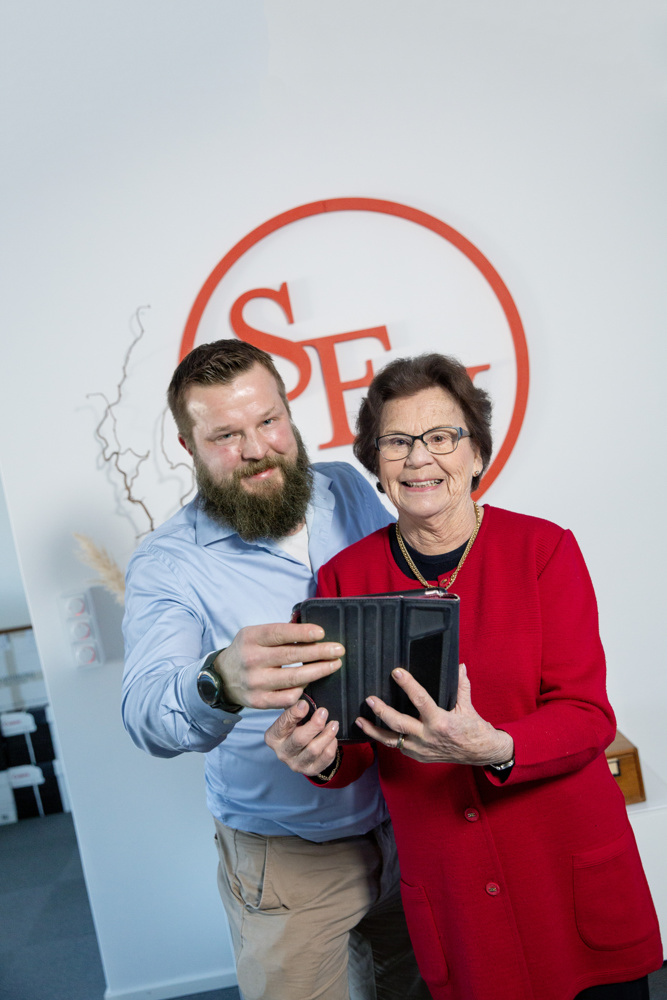 På bilden ser man en medelålders man vid namn Pasi Holmberg och en äldre dam vid namn Märta Merne Pettersson. De poserar med en dator i händerna, tittar in i kameran och skrattar. I bakgrunden ser man en ljus vägg och en logo där det står SFV i rött. Det står för Svenska folkskolans vänner. Mannen har brunt skägg och hår, ljusblå skjorta och beige byxor. Kvinnan har en röd kavaj med en svart blus under och en guldkedja runt halsen. Hon har glasögon med blå metallbågar och brunt kort vågigt hår.