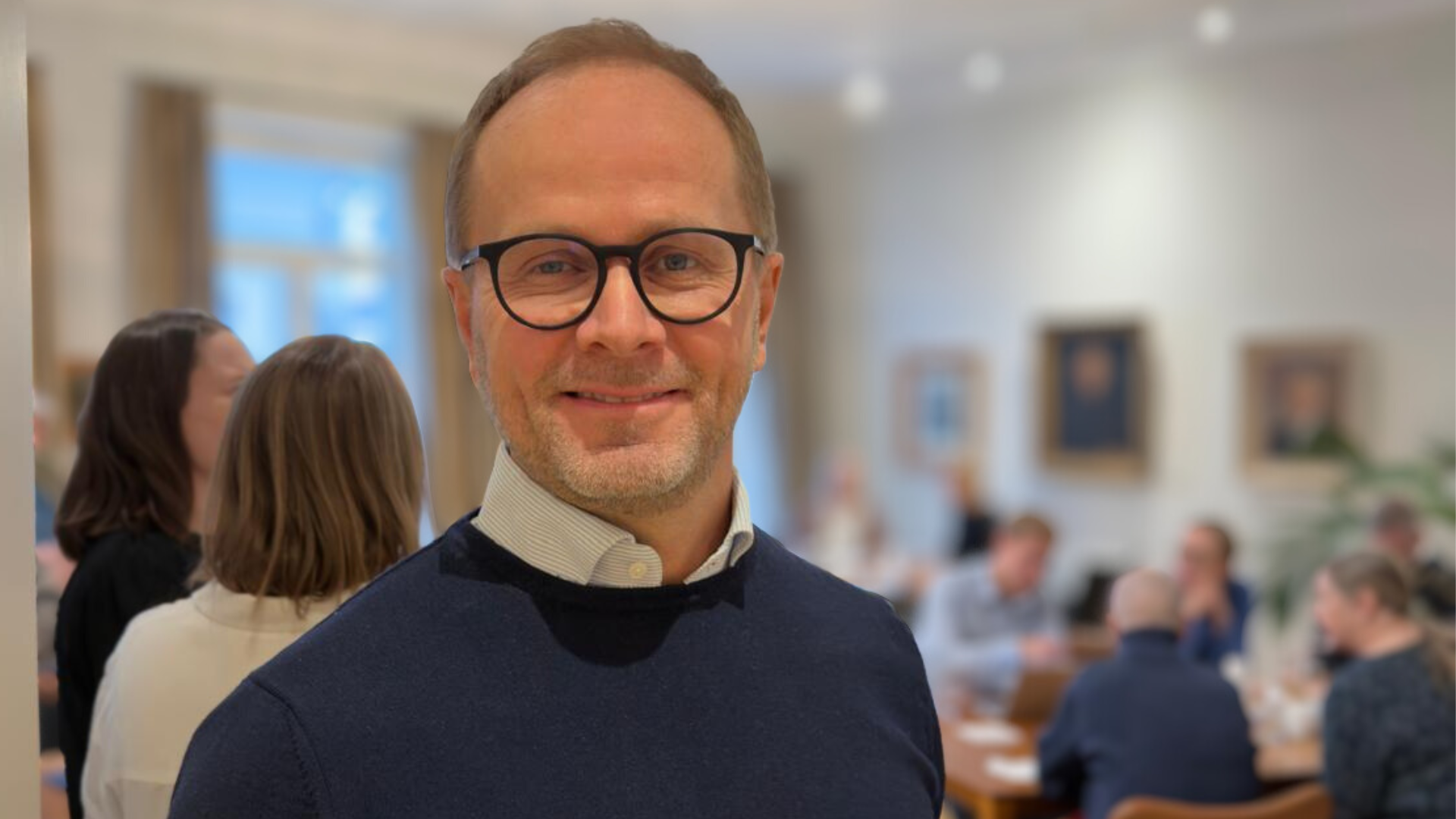 Bilden föreställer kanslichef Johan Aura i marinblå pullover och runda glasögon, leende mot kameran. Han står i dörröppningen till ett folkfyllt kontorsutrymme. 