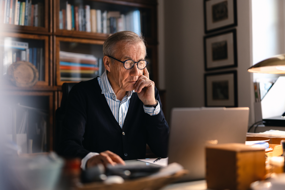 På bilden syns en äldre man vid namn Sixten Korkman. Han sitter vid ett skrivbord i trä och bakom honom ser man suddigt bokhyllor med glasdörrar. Han sitter med ena handen i hakan och tittar ner på  sin dator. På datorn ligger ett papper och en penna.