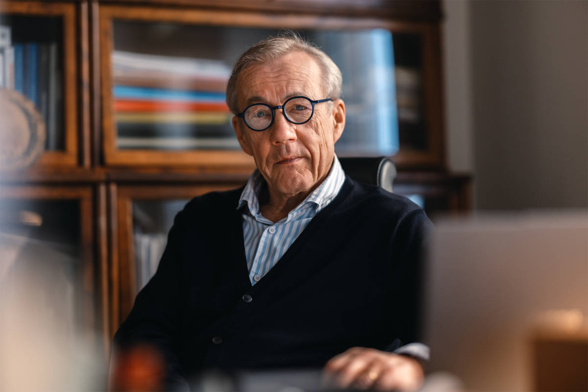 På bilden syns en äldre man vid namn Sixten Korkman med grått hos och runda glasögon som sitter vid ett skrivbord. I bakgrunden ser man inglasade bokhyllor, i framgrunden suddig en bildskärm. Mannen har en vit och ljusblå skjorta under en mörkblå tröja.