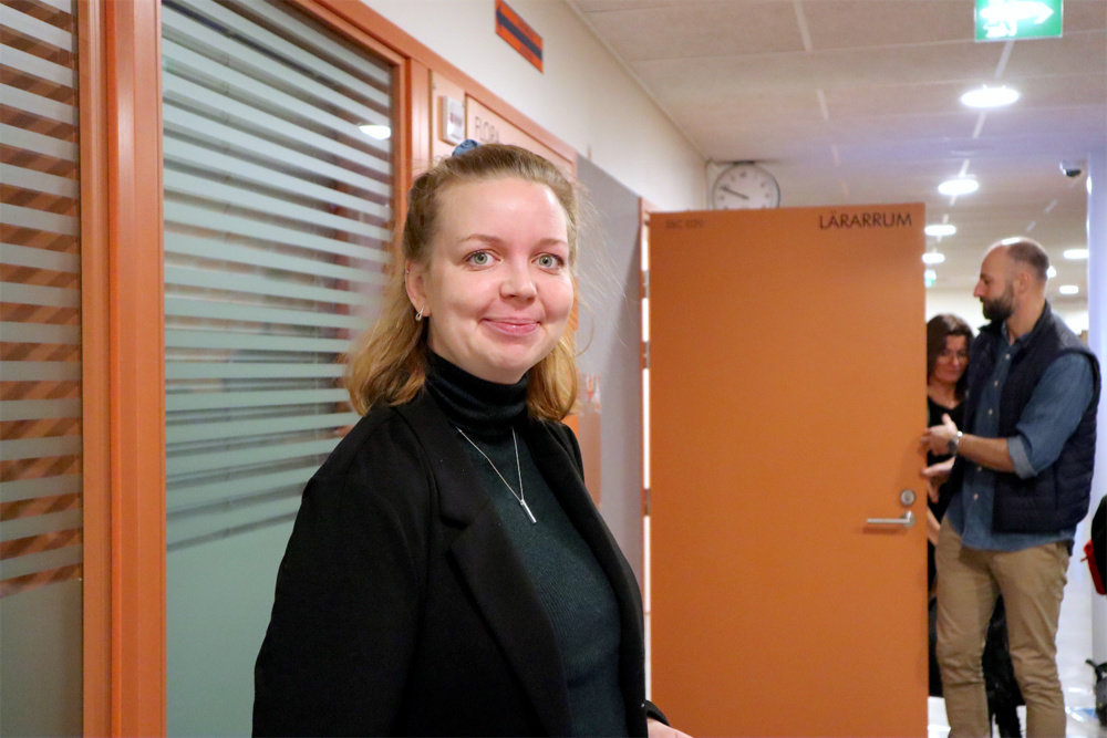 På bilden syns en medeålders kvinna med medellångt blont hår som är uppsatt upptill på huvudet och löst i nacken. Hon är klä'dd i en svart poloträja. Hon tittar in i kameran med ett litet leende. I bakgrunden ser man orangea dörrar och vita väggar. Från en av dörrarna kommer det ut en man och flera elever.