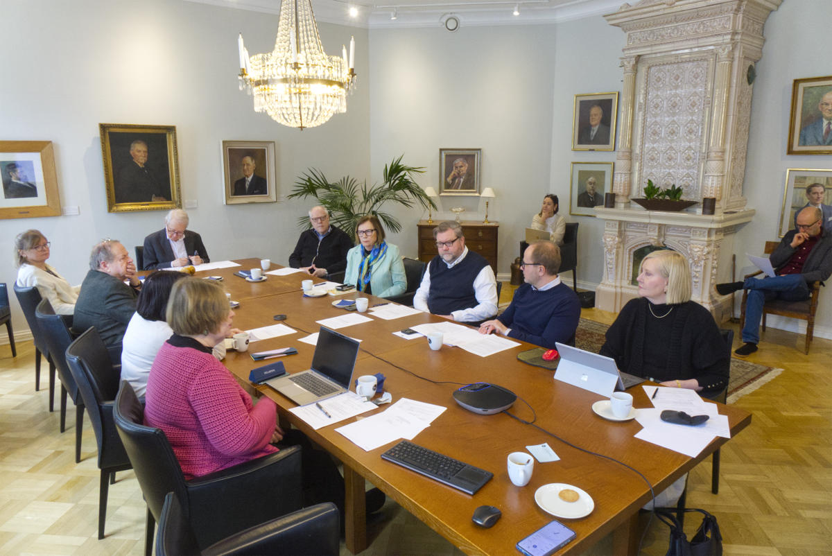 På bilden syns ett mötesrum med ett mörkt bord och svarta stolar. I taket finns en kristallkrona och på bortre väggen finns en  kakelugn i vitt, guld och rosa. Det sitter två personer på stolar bredvid kakelugnen, resten av personerna sitter vid det långa bordet. Väggarna är ljusa och det finns portsätt av mest män och ett par kvinnor upphängda längs väggarna. Det finns papper och kaffekoppar samt fat på bordet och det är ett möte på gång. Det är Understödsföreningen för svenska folkskolans vänner som har sitt årsmöte.