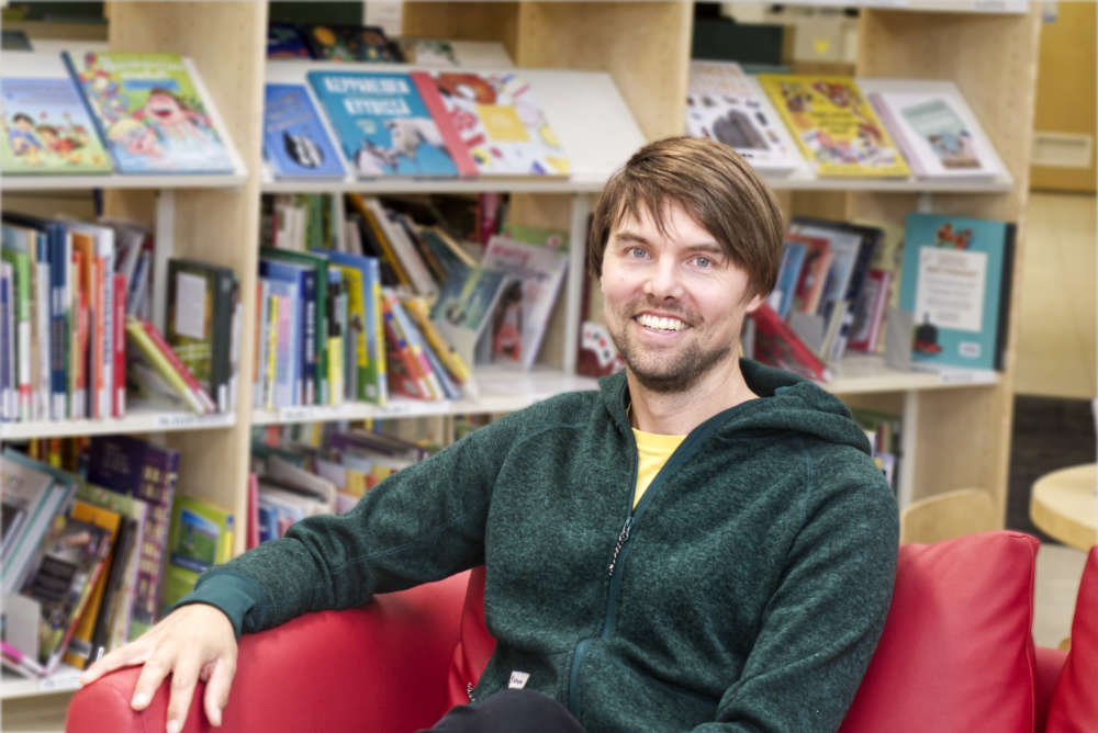 Erik BErglund ler stort mot kameran, Han sitter i en röd soffa i ett bibliotek, avdelningen är tydligt barnböcker.