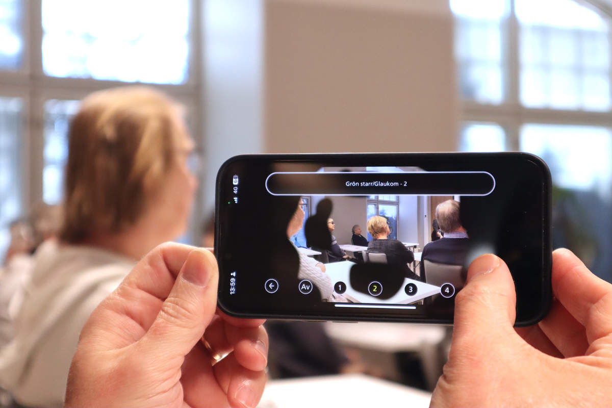 Två händer håller upp en telefon som tar in en bild från föreläsningssalen. Appen Synsimulator visar hur grön starr påverkar synen.