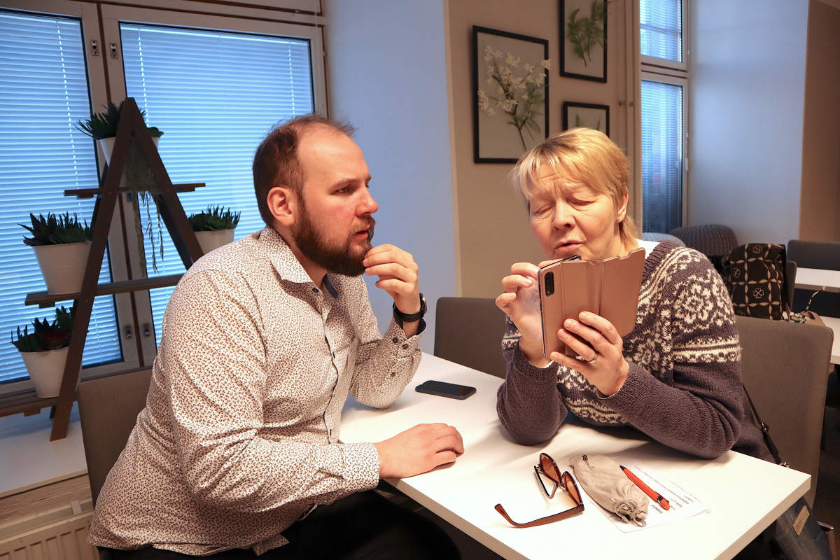 Andreas Höglund och Benita Kivimäki sitter i ett föreläsningsrumm vid vita bord. Benita håller upp sin telefon och klickar på skärmen. Andreas förklarar.