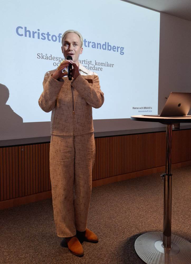 På bilden syns en komiker vid namn Christoffer Strandberg. Hans står framför en vit skärm där delar av hans namn syns i blåa bokstäver. Runt skärmen är det bruna väggar. Han är blond och på bilden ser man att han pratar med öppen mun i en mikrofon. Han är klädd i en beige kostym och har bruna skor.