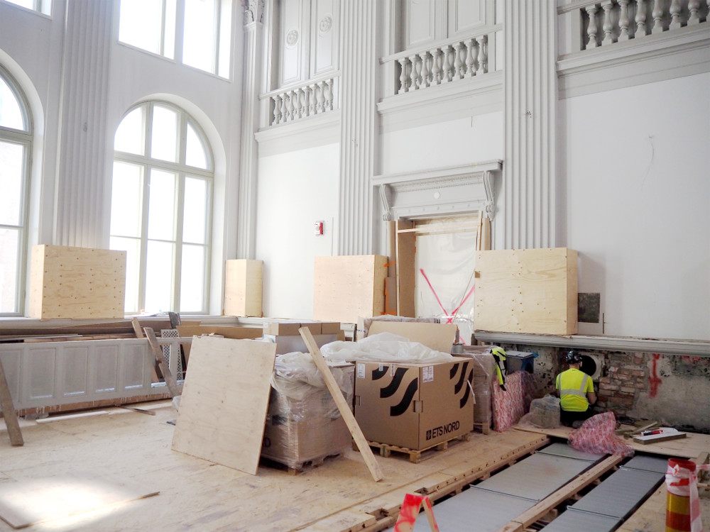 På bilden syns en sal med höga vita utsirade väggar. Det pågår en omfattande renovering och på golvet finns dels balar av byggmaterial och golvet har rivits upp. En hantverkare i neongul väst fixar något vid ena långväggen.