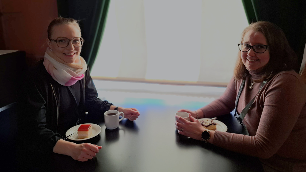 På bilden syns två kvinnor i yngre medelåldern som sitter vid ett svart bord med vita kaffekoppar framför sig. De har också varsitt kakfat med en kakbit. Kvinnan till vänster har stora fyrkantiga glasögoin och ljusbrunt hår tillbakadraget i en svans. Hon har en fluffig scarf i vitt rosa och persikofärg runt halsen och svarta kläder. HOn tittar in i kameran och ler. Kvinnan till vänster har brunt axellångt hår som är utslaget. HOn har mörka glasögin och en lingonrosa polotröja. HOn ser in i kameran och ler. I bakgrunden ser man gröna sammetsgardiner och ettt fönster med en vit rullgardin för. Foto: Sanna Mandelin