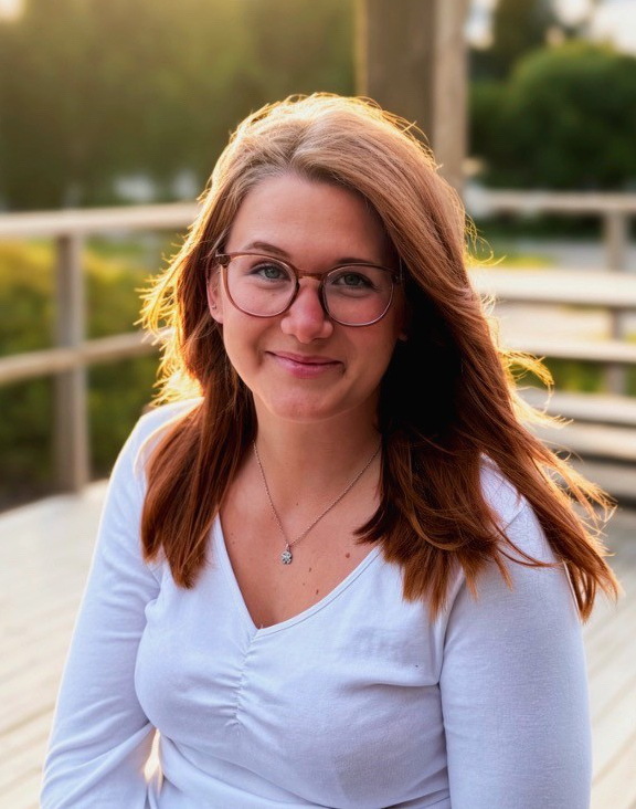 På bilden finns en ung kvinna som står på en terrass med växtlighet i bakgrunden,. Hon har långt rödbrunt hår som är löst. Hon har stora bruna glasögon, tittar rakt in i kameran och ler med stängd mun. Hon är klädd i en vit blus.