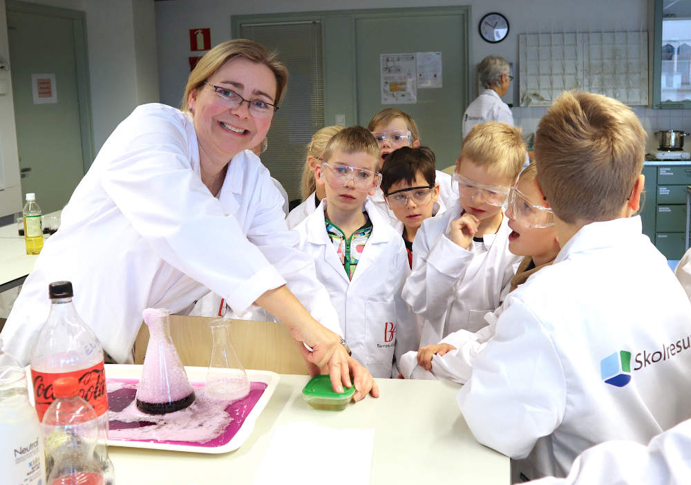 Annika Norrgård och barnen har labbrockar och skyddsglasögon på sig och har samlats kring ett labb-bord för att experimentera. 