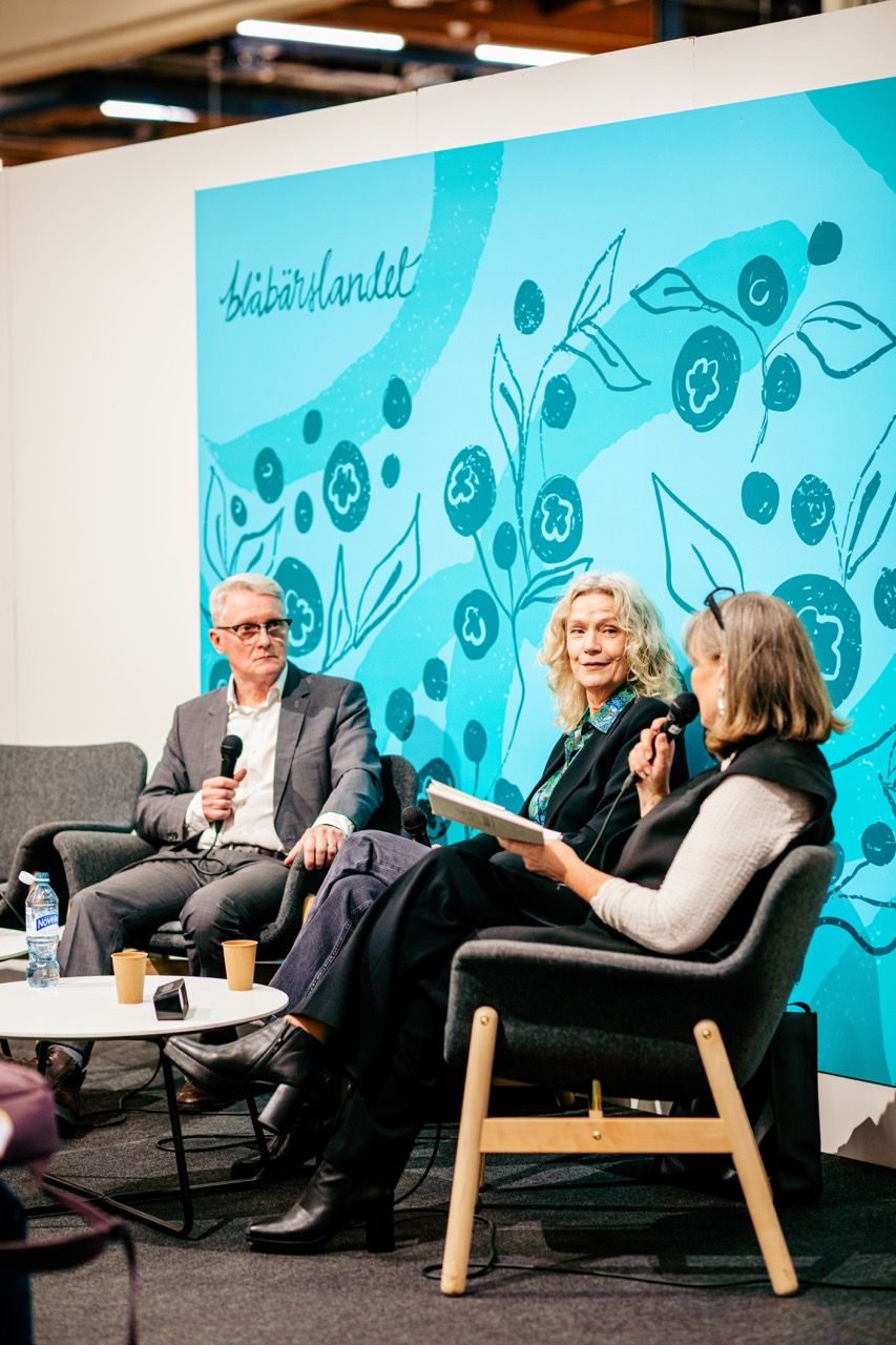 På bilden syns tre personer som sitter på en scen i gråa fåtöljer framför en väggmålning i turkost, där det står Blåbärslandet. Mannen längst till vänster är vithårig och har glasögon med tjocka svarta kalmar överst. Han är klädd i grå kostym med vit skjorta. Han håller i en mikrofon och tittar åt sidan. Kvinnan i mitten har lockigt gråblont hår ner till axlarna, en skjortblus som är mönstrad i turkost och en svart kavaj och jeans. Också hon håller en mikrofon. Kvinnan längst till höger har hår som är grått och brunt, rakt och axellångt. Hon har en vit tröja, en svart väst och svarta byxor.