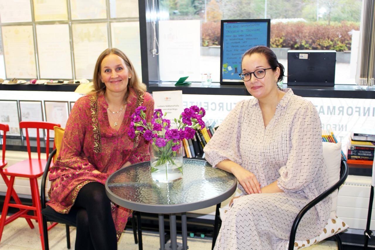 På bilden syns två kvinnor i medelåldern som sitter vid ett glasbord. Kvinnan till vänster har en röd tunika och mellanblont hår. Hon tittar in i kameran och ser glad ut. Kvinnan till höger ser allvarlig ut. Hon har mörkt hår i en knut i nacken, glasögon och en ljus lång klänning med långa ärmar.