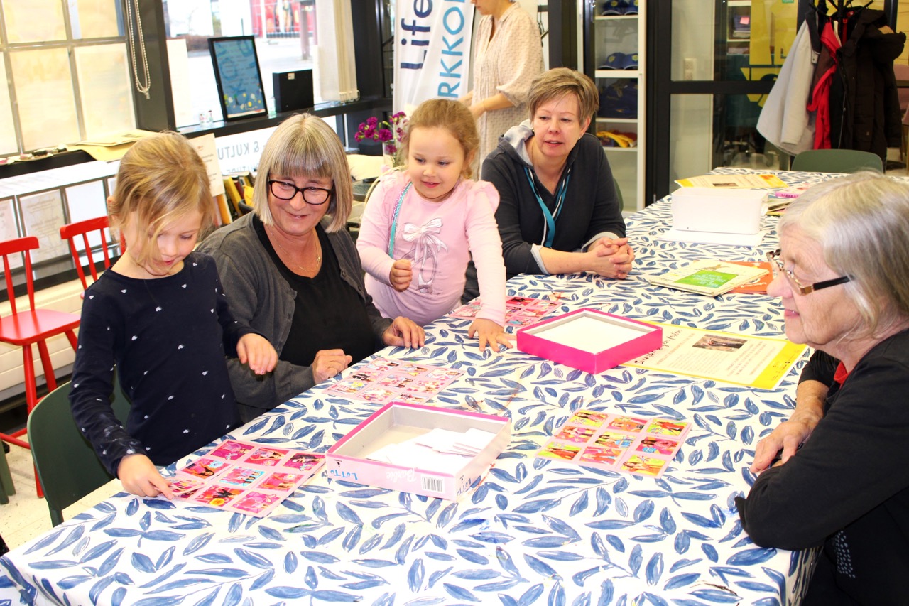 På bilden syns tre ålder damer och två småflickor som sitter kring ett bord med en uk i ljusblått och vitt. De spelar ett spel som går i rosa.