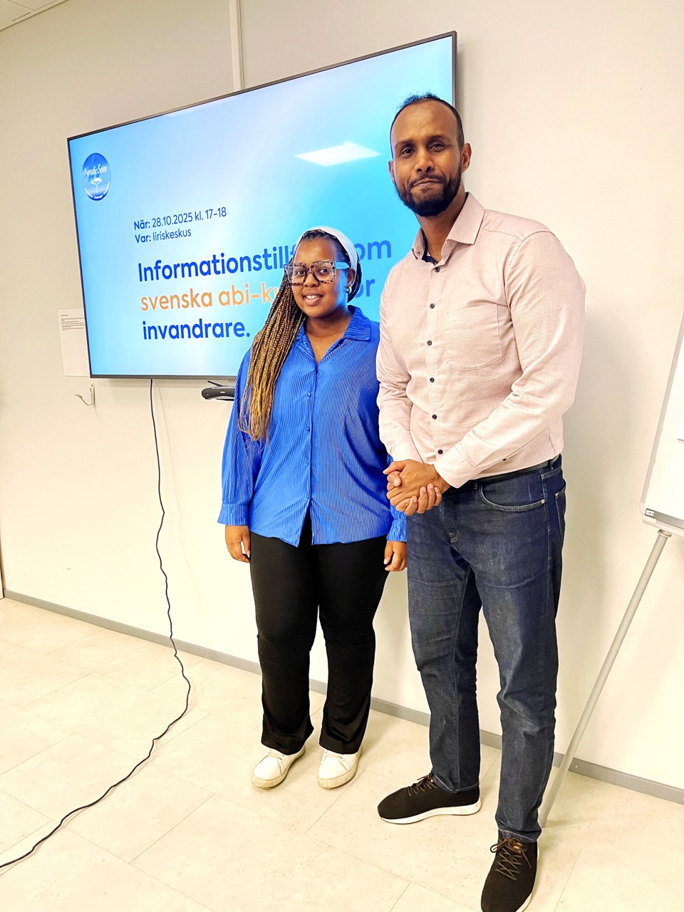 På bilden syns två mörkhyade personer. Till vänster står en ganska kort kvinna, med stora glasögon, vitt diadem i håret och långa flätor över ena axeln. HOn har en klarblå skjortblus, svarta byxor och vita skor. Till höger syns en lång man  men kort helskägg, rosa skjortblus, blåa jeans och bruna skor. De står framför en vit vägg i ett klassrum och en blå skärm syns bakom dem.