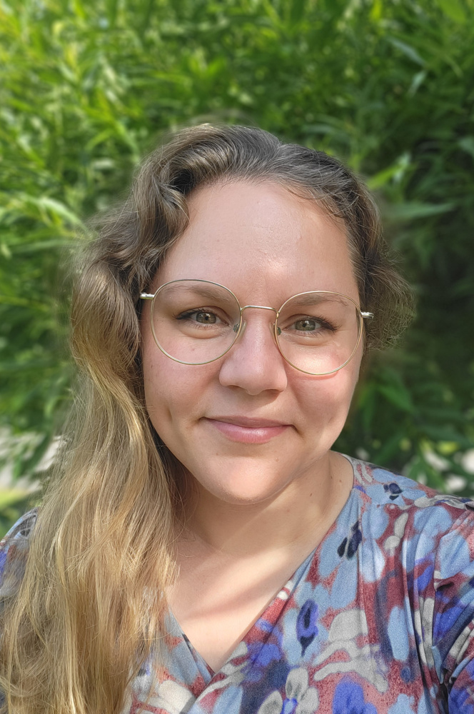 På bilden syns en medelålders kvinna som är fotograferad utomhus. I bakgrunden syns grön växtlighet. Hon har långt ljust hår som hänger över ena axeln. Hon har runda metallfärgade glasögon och tittar rakt in i kameran och ler med stängd mun. HOn är klädd i en blommig blus med röda, blåa och vita blommor.