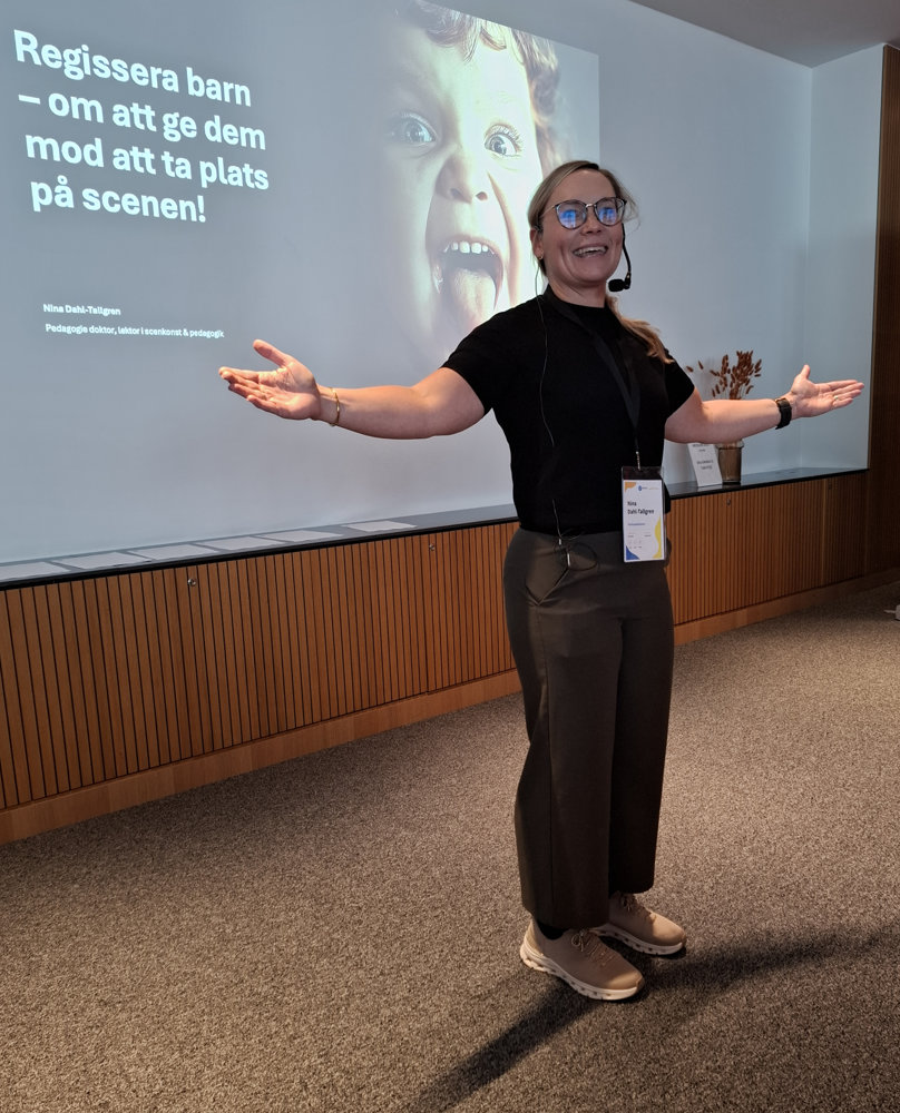 På bilden syns en föreläsare vid namn Nina Dahl-Tallberg som talar om att ge barn mod att uppträda. Hon har mellanblont hår som är fast i nacken. Hon har glasögon och är klädd i en svart t-shirt och bruna byxor. Bakom henne ser man en skärm som snett har texten Regissera barn - om att ge dem mod att våga ta plats på scenen