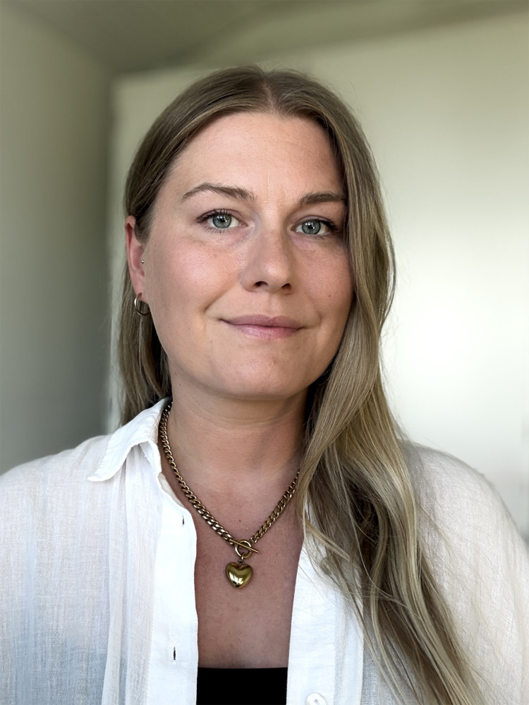På bilden syns en medelålders kvinna fotad mot en ljusgrå bakgrund. Hon har mellanblont hår som hänger över en axel. Hon tittar rakt in i kameran och ser allvarlig ut. Hon är klädd i en vit skjortblus ovanpå en svart topp.
