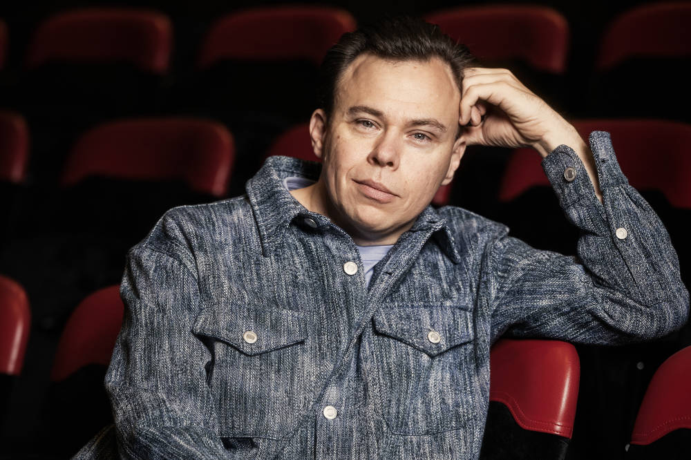 Carl Knif lutar sig mot ena armen. Han sitter ensam på en stolsrad i en teatersalong. 