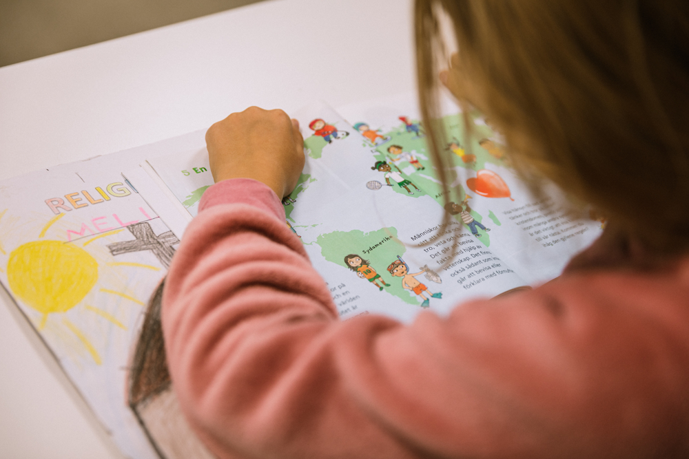 På bilden syns en flicka bakifrån. Hon har mellanblont hår och sitter och skriver i en färgglad bok med lite text. Hon sitter vid ett vitt bord.