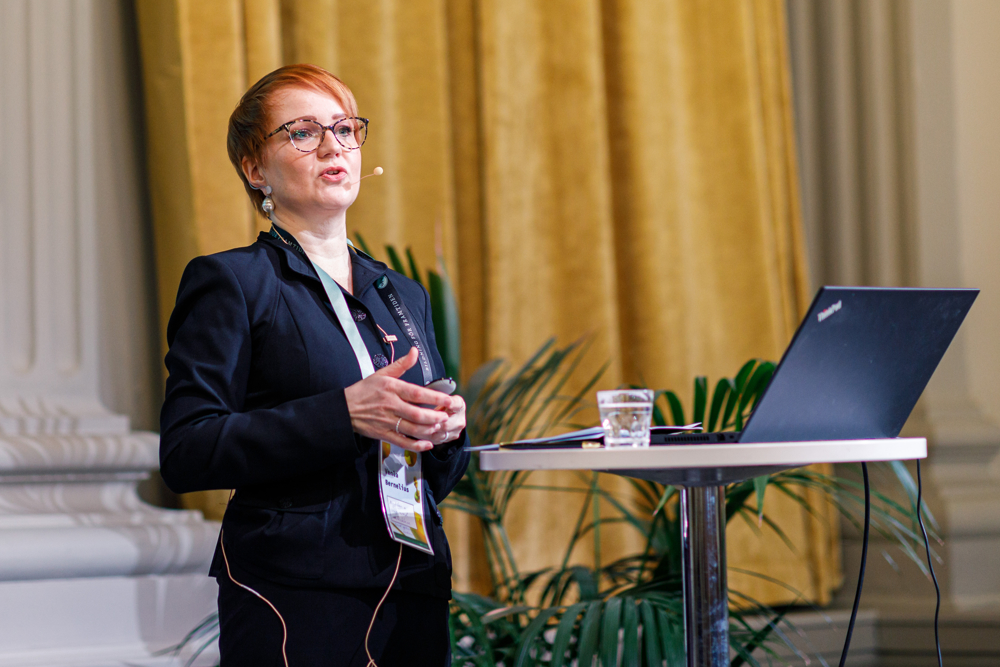 På bilden syns en ung kvinna med kort rött hår som står vid ett talarpodium. HOn har mikrofon och sändare på sig och håller ett tal. Kvinnan heter Venla Bernelius och är specislsakkunnig på Undervisnings- och kulturministeriet och uttalar sig om framtidensrapporten om grundskolan. På bilden är hon klädd i en mörkblå kavaj och en mörkblå kjol. Hon har stora glasögon med brunfläckiga bågar. I bakgrunden ser man vita pelar och ett gult draperi.